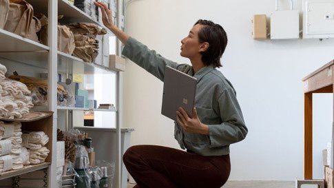 A woman holds Surface Pro 7  while reaching up to a bookshelf